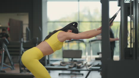 A-young-Hispanic-woman-in-a-yellow-tracksuit-performs-an-exercise-in-a-crossover-pulls-a-rope-from-above-to-train-the-back-and-shoulders.-Brunette-woman-trains-back-and-shoulders-in-the-gym